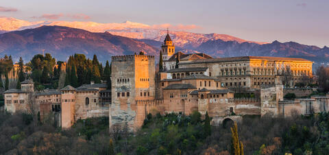 A view of the Alhambra palace in Granada, illuminated by warm sunset light, with snow-capped mountains and lush trees in the background