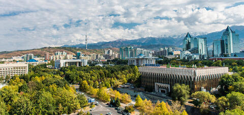  A panoramic view of Almaty, Kazakhstan, showing modern buildings surrounded by green spaces, with the snow-capped mountains in the background