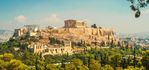 A view of the Acropolis in Athens, Greece, featuring the Parthenon atop a rocky hill surrounded by greenery, with the cityscape visible in the background