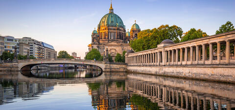 Berlin Cathedral with its green dome is reflected in the waters of the Spree River at sunset, with a bridge and colonnade in the foreground