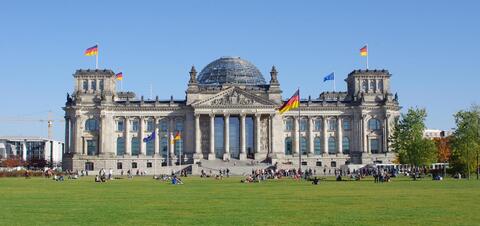 Tourists relax on the lawn in front of the historic Reichstag Building in Berlin, Germany, with German flags flying and the glass dome visible atop the structure