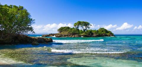 A small, tree-covered island off the coast of a beach in Santiago, surrounded by clear turquoise waters and rocky shores