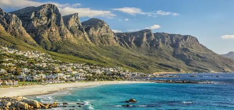 An aerial view of Cape Town, South Africa, showing the coastal city nestled beside rugged mountains, with sandy beaches and the blue waters of the Atlantic Ocean