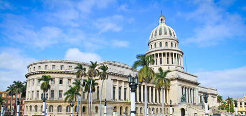 The grand El Capitolio Nacional building in Havana, Cuba, featuring a large domed roof, neoclassical architecture, and palm trees lining the entrance.