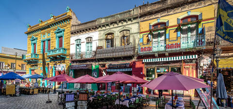 Outdoor cafe tables with umbrellas set against a backdrop of colorful historic buildings in the La Boca neighborhood of Buenos Aires, featuring bright yellow, blue, and red facades with intricate balconies