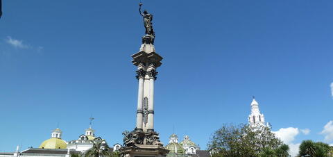 The Independence Monument in Plaza Grande, Quito, featuring a tall column with a statue honoring the heroes of Ecuador's independence, with ornate domes and historic buildings in the background.