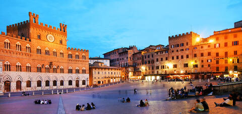 A twilight view of Piazza del Campo in Siena, with small groups of people sitting in the large piazza, surrounded by the illuminated red brick Palazzo Pubblico