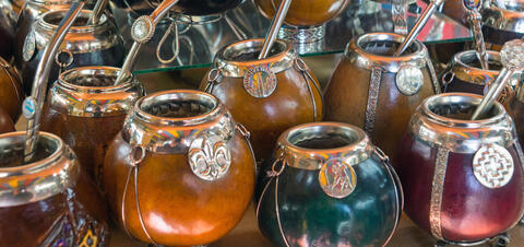 An assortment of traditional Argentine calabash gourd mate cups, each adorned with silver decorations and metal straws (bombillas), displayed together in a vibrant market setting