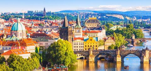 A panoramic view of Prague featuring the iconic green-domed buildings, gothic towers, and the historic Charles Bridge crossing the Vltava River