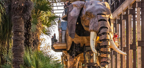 A large mechanical elephant carrying passengers through Les Machines de l'Île in Nantes, France, surrounded by trees and industrial structures