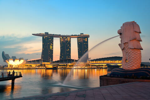 A view of Singapore's iconic Merlion statue with water spouting from its mouth, overlooking the Marina Bay Sands hotel and skyline at dusk, with the bay reflecting the evening lights.
