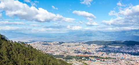 A panoramic view of Quito, Ecuador, with the city's buildings spreading across the valley, surrounded by green hills and distant mountains
