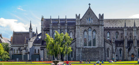 A large stone cathedral with tall spires and arched windows, set against a blue sky, with a park and people relaxing in front