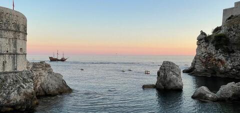 A scenic view of a fort by the sea with a sailing ship and kayakers on the water, framed by rocky cliffs