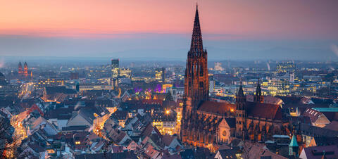 A panoramic view of Freiburg, Germany, at twilight, featuring the illuminated spire of the Freiburg Minster and the softly glowing city skyline 