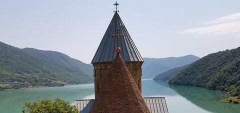 The pointed towers of Ananuri Castle rise in front of a serene river and surrounding green mountains
