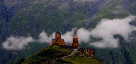Gergeti Trinity Church sits atop a green hill with two pointed towers, surrounded by misty clouds and towering mountains in the background