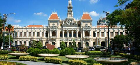 The People's Committee Building in Ho Chi Minh City, a grand French colonial structure with arched windows, red-tiled roofs, and an ornate central clock tower,with green trees and gardens in the foreground