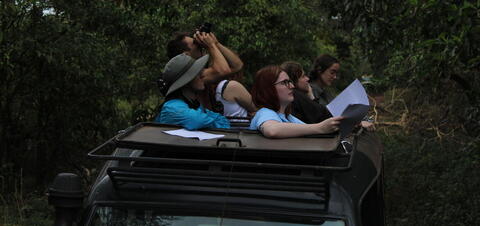 Students in an open top vehicle in the forest using binoculars and referencing their notebooks to identify Olive Baboon Behaviors at Lake Manyara