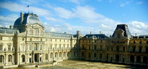 The Louvre Museum, a grand and ornate complex of stone buildings with intricate facades, bathed in sunlight under a clear blue sky