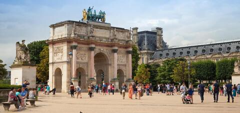 Tourists walk near the Arc de Triomphe du Carrousel, a triumphal arch in front of the Louvre Museum, with sculptures and ornate architecture in the background
