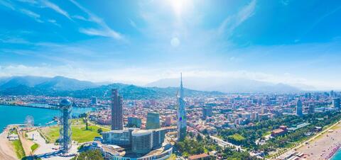 A panoramic view of Batumi, Georgia, featuring colorful buildings with ornate porches and red tile roofs sprawling up a mountainside, with modern skyscrapers and the coastline in the foreground