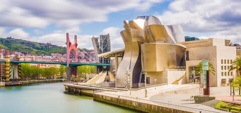 The Guggenheim Museum in Bilbao, featuring its unique, curving metallic architecture beside the river, with the red La Salve Bridge and the city in the background 