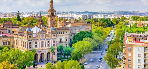 A scenic view of Seville, Spain, featuring the historic General Captaincy building with its ornate architecture, surrounded by lush green trees and a busy city street