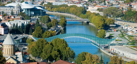 A view of Tbilisi, Georgia, featuring the modern glass Bridge of Peace spanning the Kura River, with trees lining the riverbanks and contemporary architecture in the background