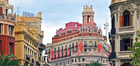 A building with a red facade and the words "banco de Valencia" stand at the end of a street lined with other ornate buildlings.