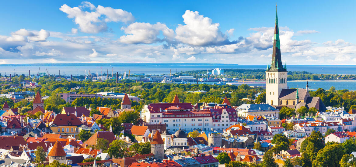 A scenic view of Tallinn's historic red-roofed buildings and the tall spire of St. Olaf's Church, with the Baltic Sea and harbor in the background