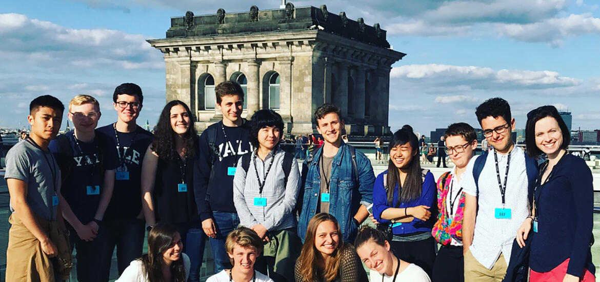 A group of smiling students stands outside in front of a bridge and a river