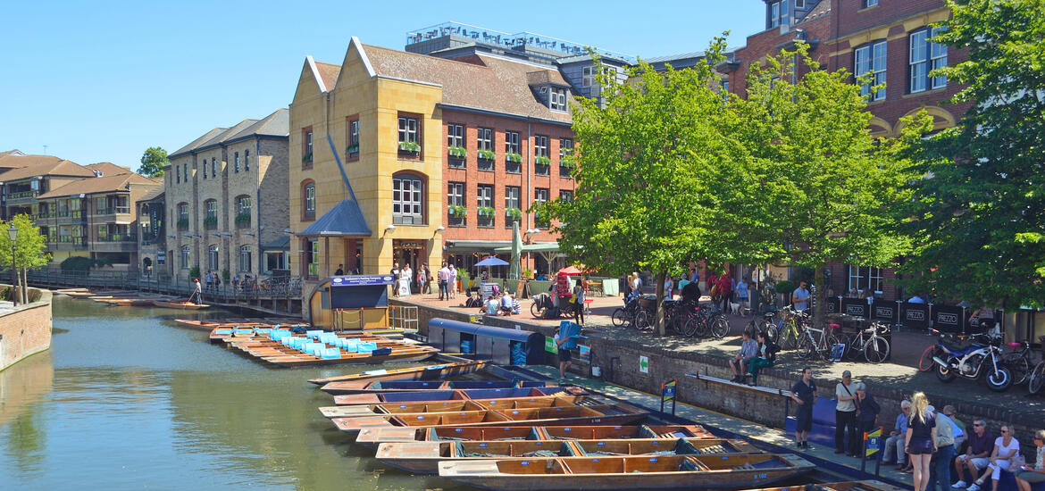 A view of the River Cam in Cambridge, England, with tourists preparing for punting near Magdalene College, surrounded by historic buildings, trees, and outdoor cafes